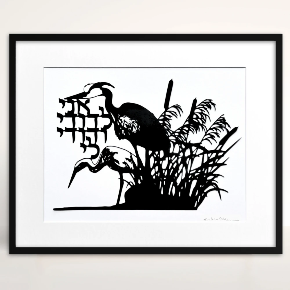 Black papercut artwork depicting two herons in a marsh setting with cattails and reeds. The artwork includes Hebrew text and is set against a white background. The herons are in silhouette.