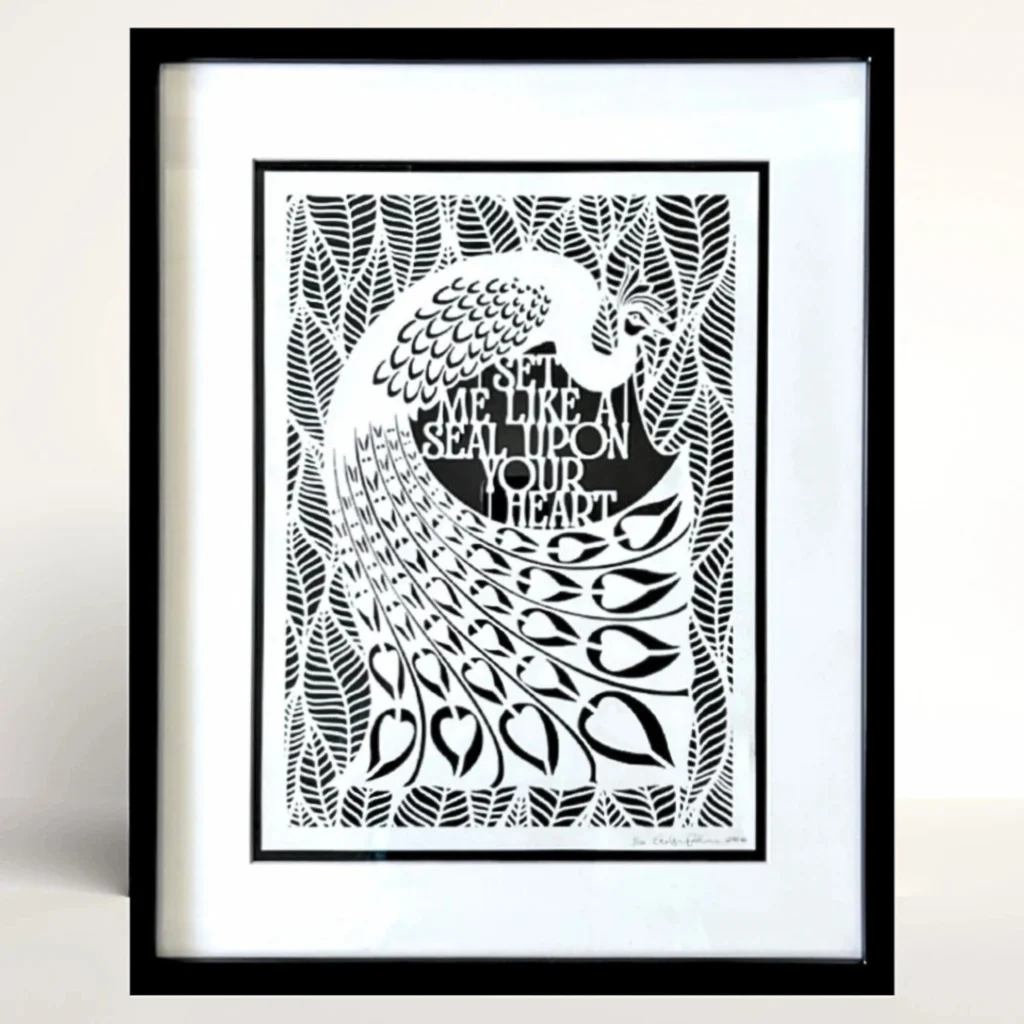 The peacock in the middle of the papercut is surrounded by broad leaves. The English text for the quote sits between the peacock's curved tail and body. By Evelyn Goldman Art