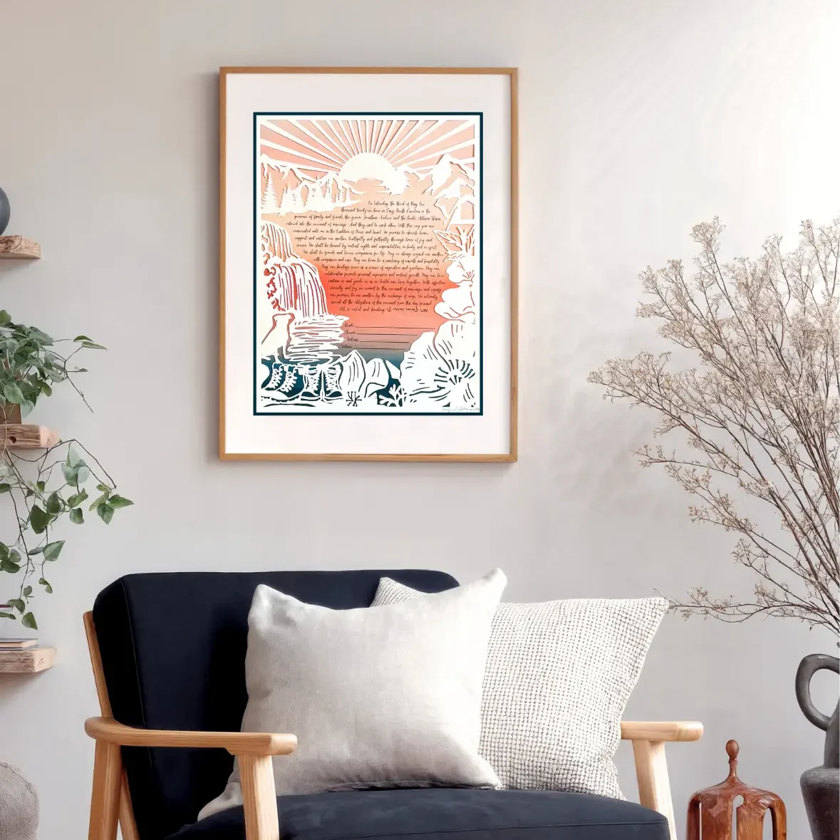 A framed paper-cut artwork ketubah depicts mountains, trees, a waterfall and river, a pair of boots, a dog, and a tent with a handwritten marriage contract at the center, beneath a sunrise. This is hanging on the wall of someone's home
