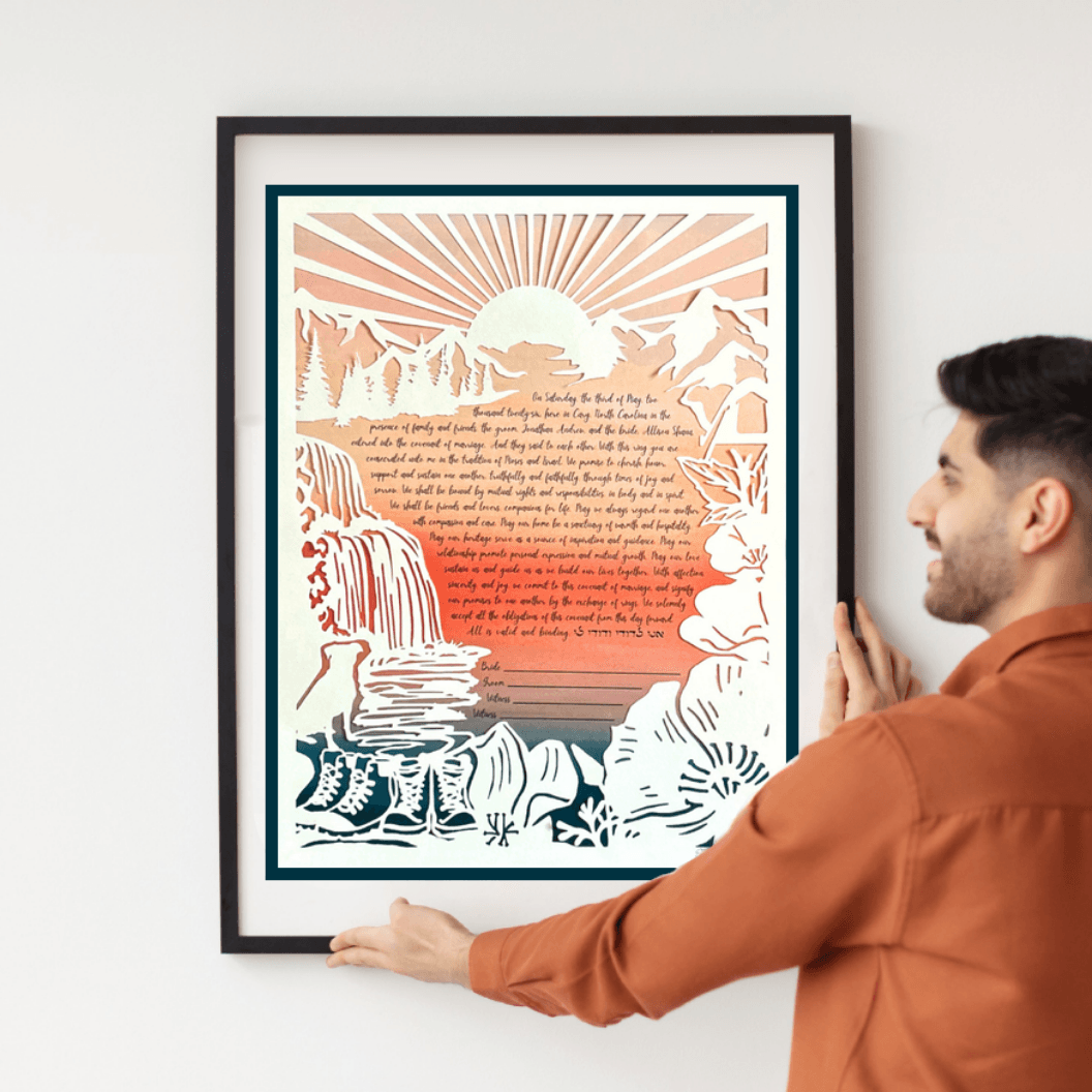 A paper-cut artwork ketubah depicts mountains, trees, a waterfall and river, a pair of boots, a dog, and a tent with a handwritten marriage contract at the center, beneath a sunrise.