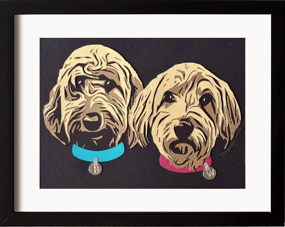 Framed layered paper cut artwork of two doodle dogs with textured fur, one wearing a blue collar and the other a pink collar, each having a tag with their initial letter on it.