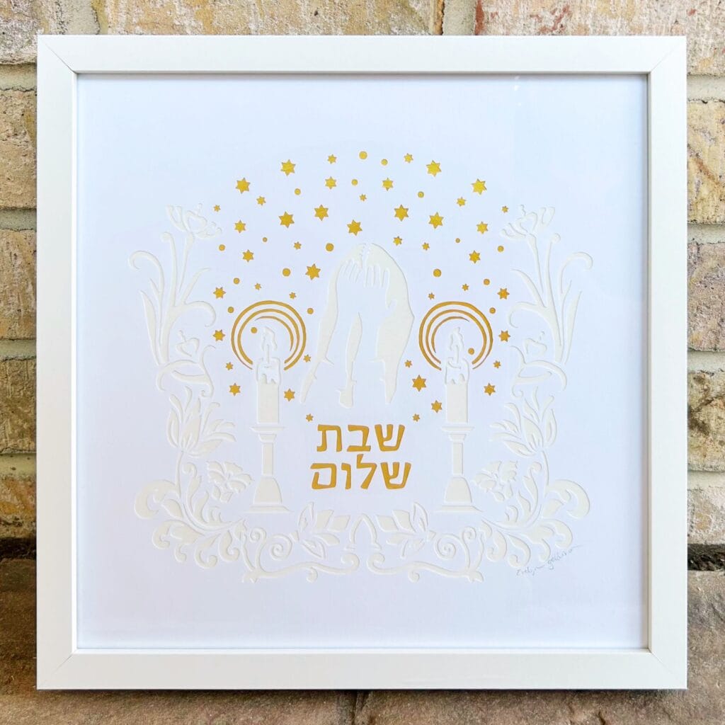 Framed paper cut art, white on white with gold stars of David and other gold details, art nouveau floral designs, Hebrew text that says Shabbat Shalom, and a woman covering her eyes and blessing the shabbat candles.