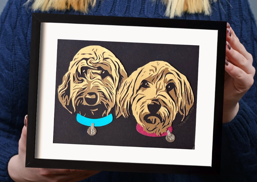 A woman holds her framed pet portraits of her two dogs. The frame is black with a white mat